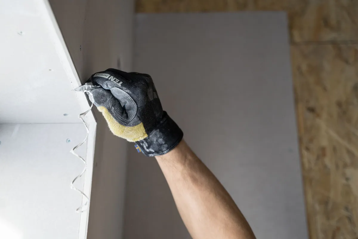 A gloved hand running a taping knife down a drywall corner bead.
