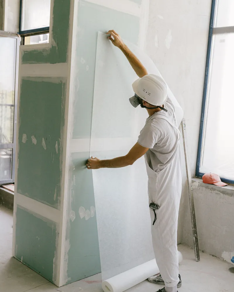 A worker lifting a sheet of drywall into place against a wall.