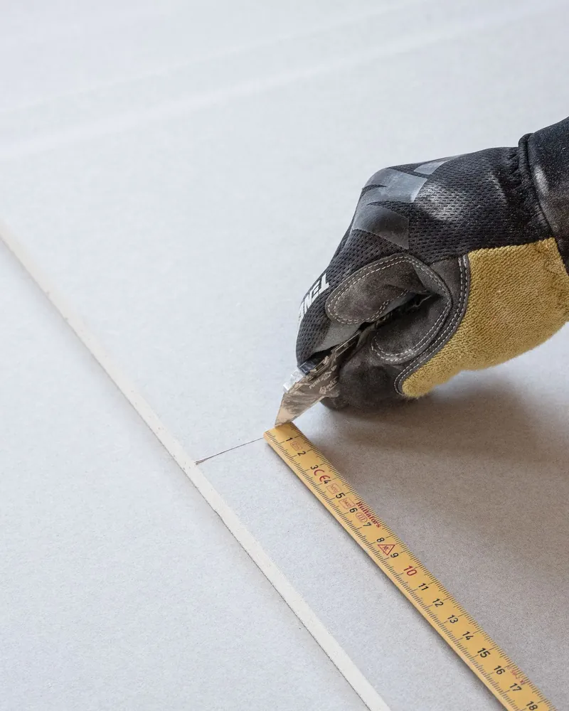 A gloved hand measuring a sheet of drywall with a tape measure.